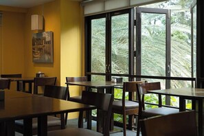 A restaurant with wooden tables and chairs, large windows with a view of palm trees, and a painting on the wall. - The Bihai Hua Hin (Hua Hin)