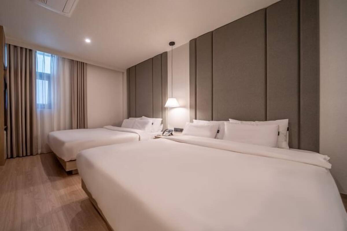 A hotel room with two beds, a large window with curtains, a wall-mounted lamp, and a wooden floor.