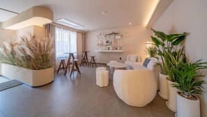 Una sala de estar moderna con sofá, otomanas, una mesita de centro y un comedor con sillas y una mesa pequeña. Hay plantas en macetas y una estantería con objetos decorativos.