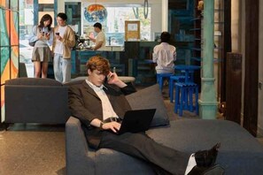 Une personne assise sur un canapé avec un ordinateur portable, un coin bar avec des tabourets, et un groupe de personnes à l’arrière-plan.