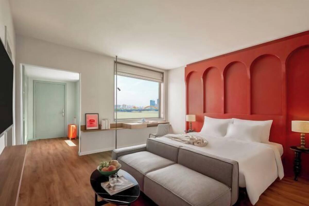 Une chambre d’hôtel avec un lit, un canapé, une petite table sur laquelle se trouve un saladier de fruits, une fenêtre donnant sur un paysage agréable et un mur d’accent de couleur rouge.