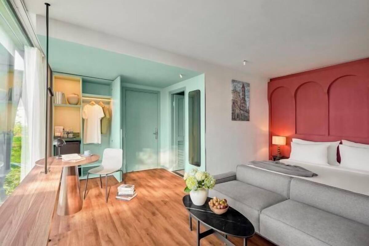 Une chambre d’hôtel moderne avec un plancher en bois, un canapé gris, une petite table sur laquelle est posé un vase de fleurs, un lit avec du linge de lit blanc et un mur d’accent rouge.