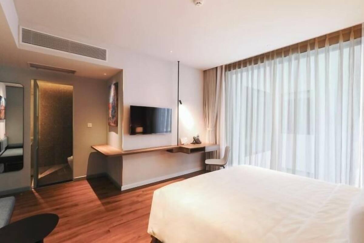 Une chambre d’hôtel moderne dotée d’un grand lit, d’un téléviseur à écran plat fixé au mur, d’un bureau avec une chaise et d’une fenêtre avec des rideaux légers.