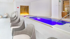 A modern indoor pool area with a long, narrow pool, seating along the sides, and a glass door leading to another room.
