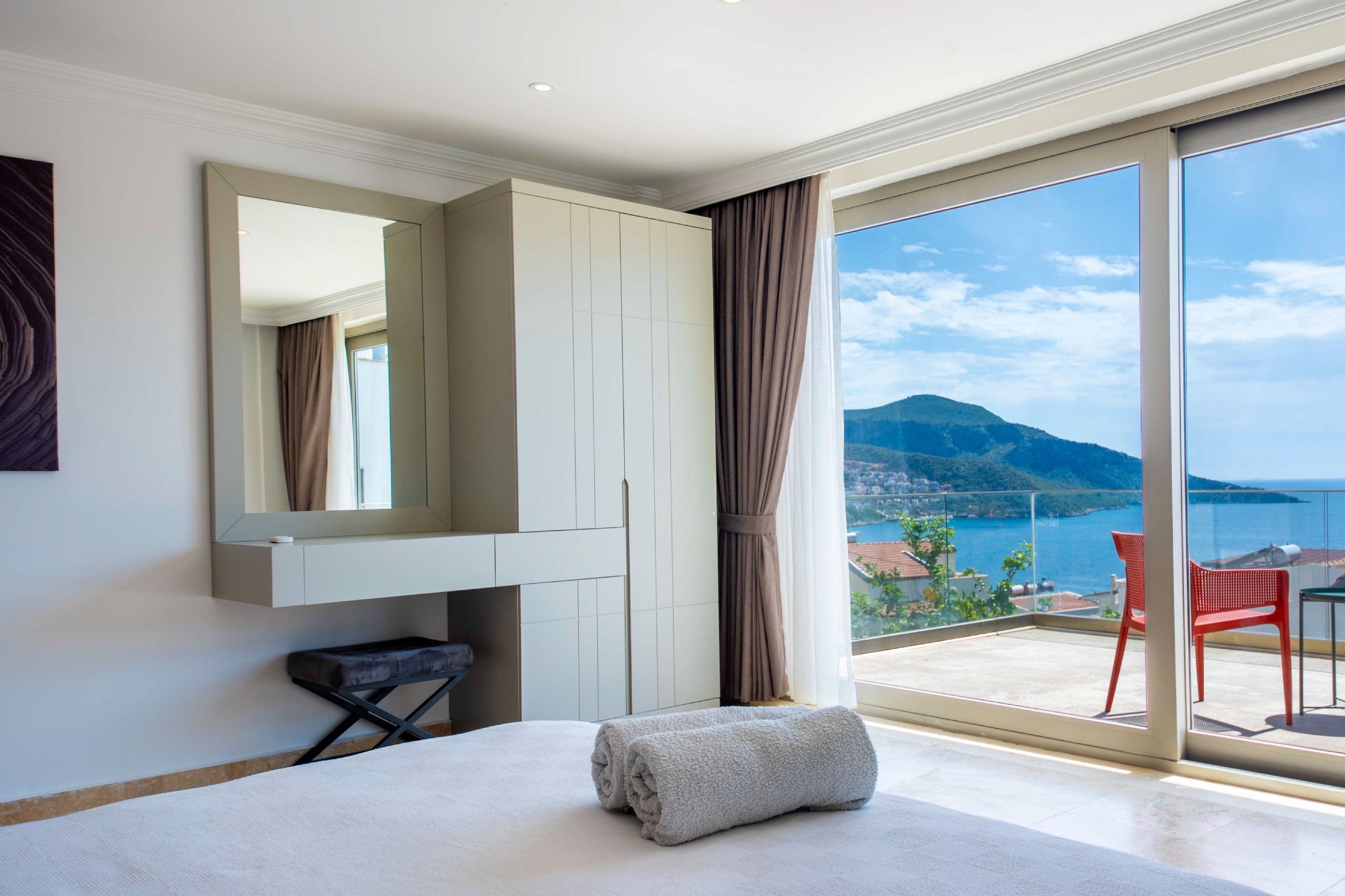 A hotel room with a bed, a chair, a large window with a view of the sea and mountains, and a wardrobe.