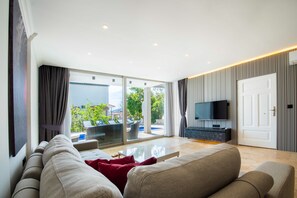A modern living room with a sofa, a flat-screen TV, and a glass door leading to an outdoor area with a pool. - Steps from Kalkan’s beach and tavernas (Kalkan Centre)