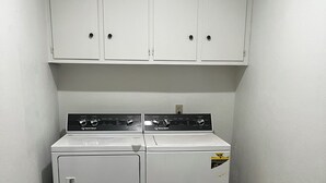 Une buanderie équipée de deux machines à laver et deux sèche-linge à chargement frontal superposés, avec des placards blancs au-dessus et un sol carrelé en pierre.