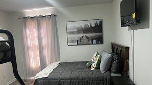 Une chambre à coucher comprenant un lit, une tête de lit, une table de chevet, une chaise, une fenêtre avec des rideaux et un téléviseur fixé au mur.