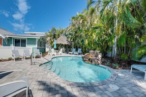 A swimming pool with lounge chairs and a thatched-roof structure, surrounded by palm trees.