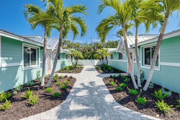 A pathway flanked by palm trees leading to a row of light green buildings with white trim.