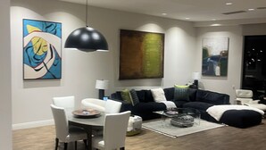 A modern living room with a dining area, featuring a black pendant light, a large abstract painting, and a dark-colored sofa with cushions. - Elegant Private 2-Bedroom Ensuite Condo with Expansive Living and Dining Areas (Rancho Mirage)