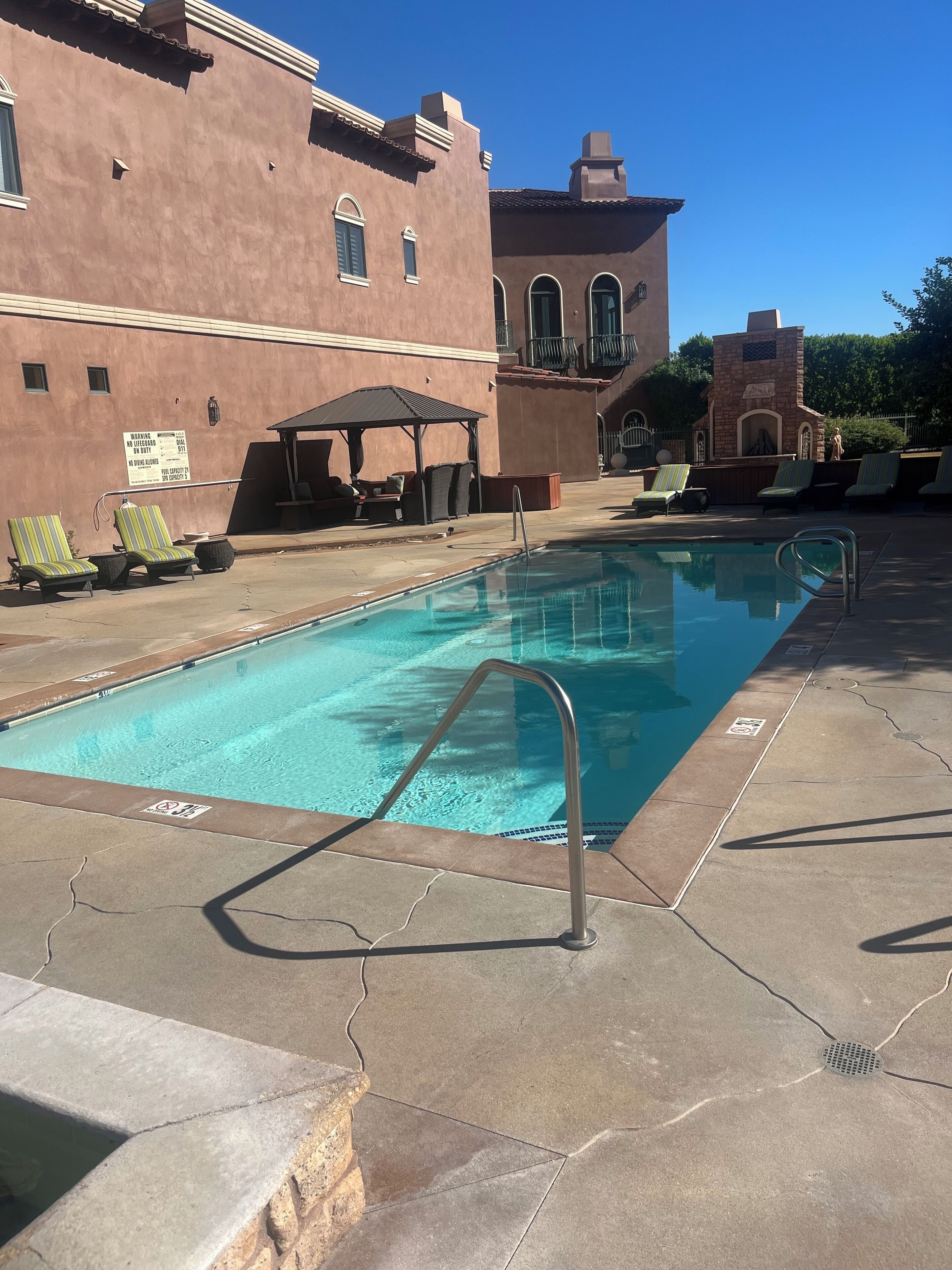 Une piscine entourée de chaises longues et d’un espace de détente couvert, le tout bordé d’un bâtiment doté d’un toit en tuiles et d’une cheminée en pierre.