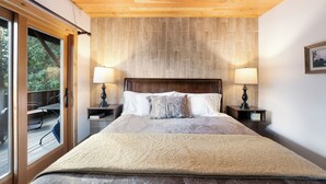 A bedroom with a wooden ceiling, a large bed with a beige quilt, two bedside tables with lamps, and a view of an outdoor deck through a sliding glass door. - Epic Group Stay | 3 Cabins w/ Views | Near Glacier NP, Skiing & Summer Adventures! (Columbia Falls)