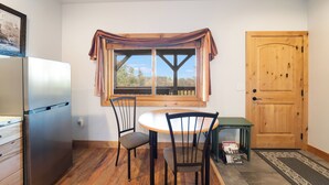 A kitchen with a wooden table, two chairs, a refrigerator, a window with a curtain, and a wooden door. - Epic Group Stay | 3 Cabins w/ Views | Near Glacier NP, Skiing & Summer Adventures! (Columbia Falls)