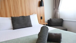 A neatly made bed with a green bedspread, two rolled towels, a wooden headboard, a bedside table, a lamp, and a window with curtains. - Noa Hotel (Auckland)