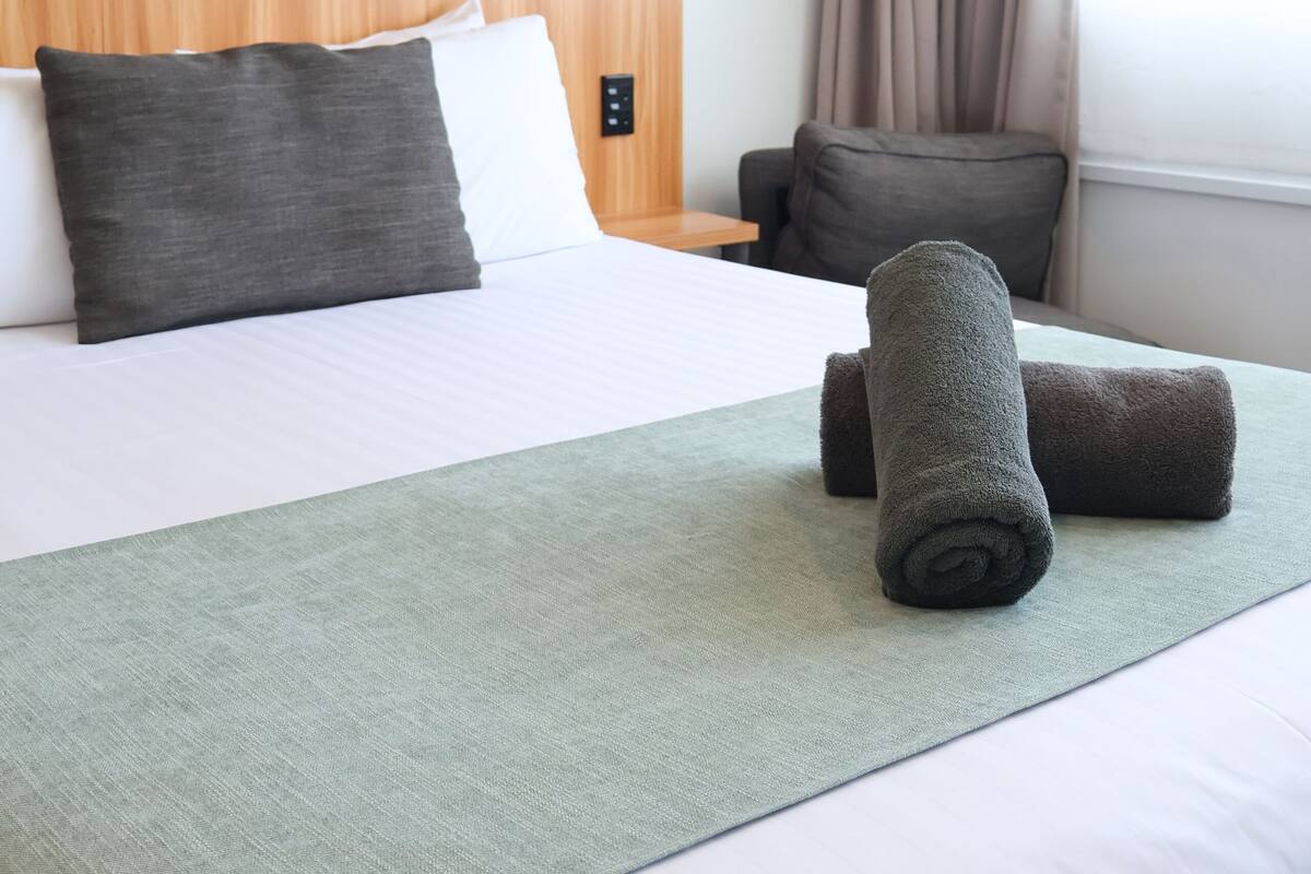 Ein Hotelzimmer mit einem hölzernen Kopfteil, einem Bett mit weißer und grauer Bettwäsche, einer grünen Tagesdecke und zwei zusammengerollten Handtüchern auf dem Bett.