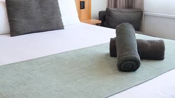 A hotel room with a wooden headboard, a bed with white and gray bedding, a green throw blanket, and two rolled towels on the bed.