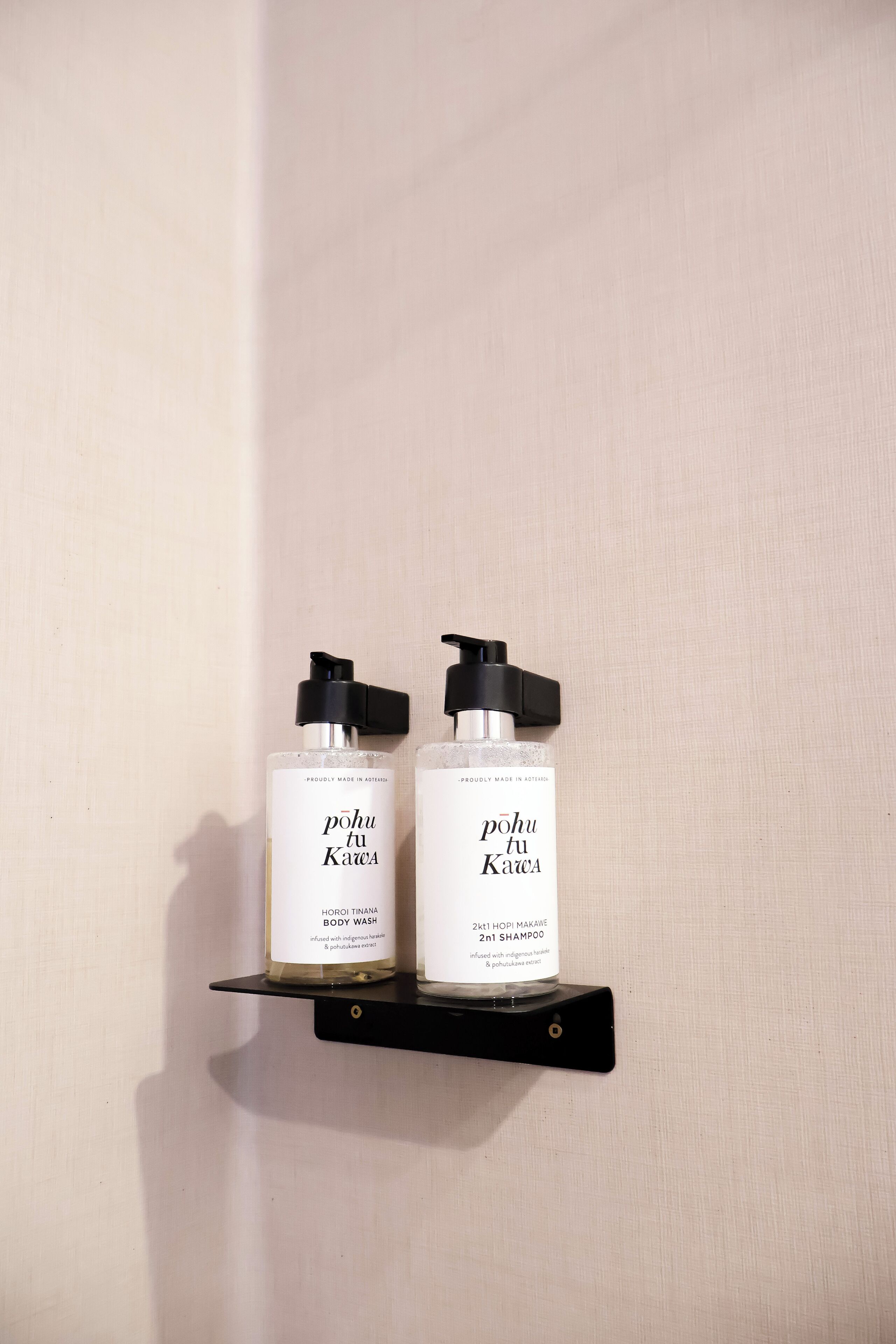 Dos botellas de jabón de cuerpo y champú sobre un estante negro frente a una pared clara.