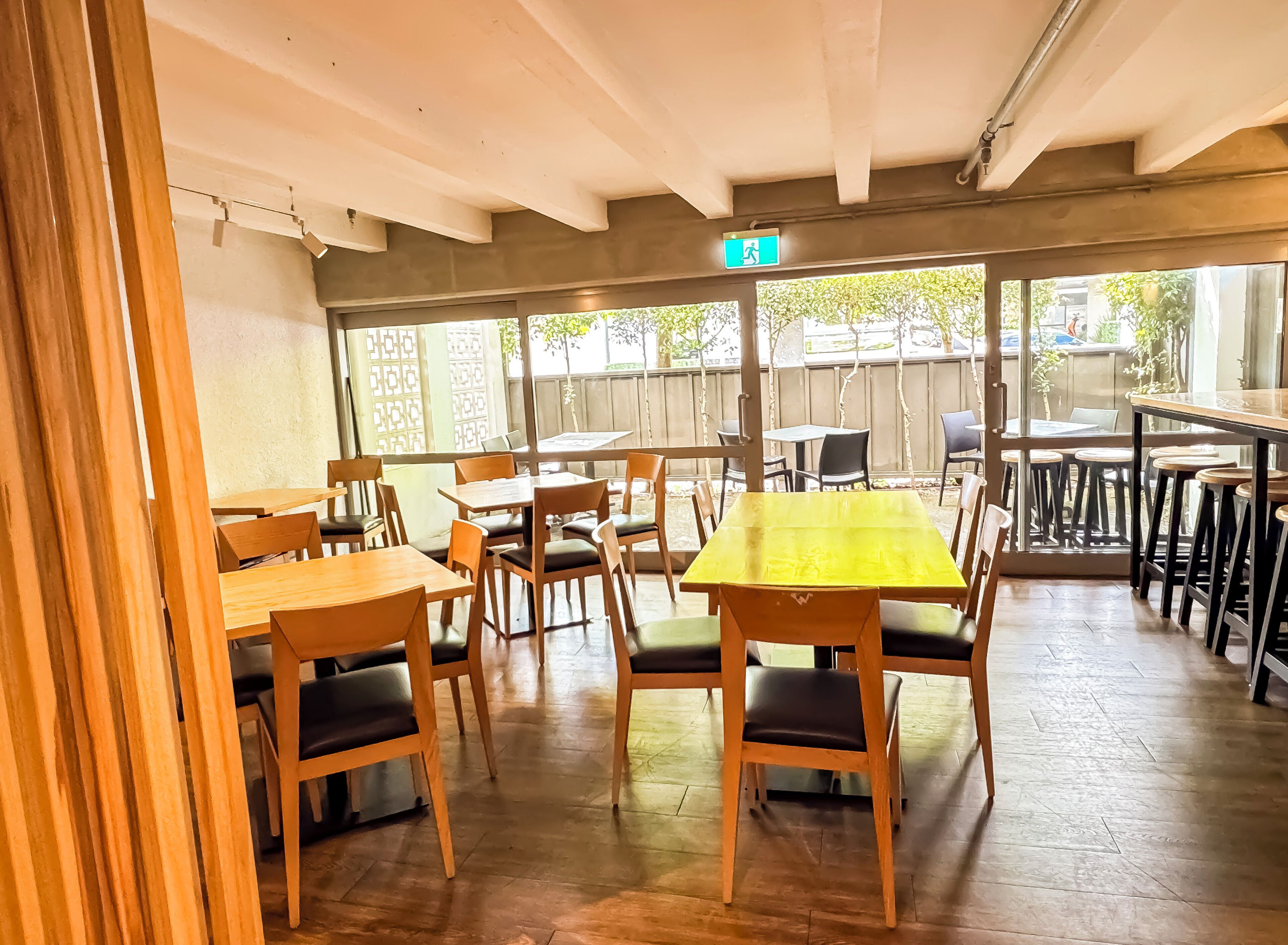 A restaurant with wooden tables and chairs, a yellow table in the center, and a view of the outdoors through large windows.