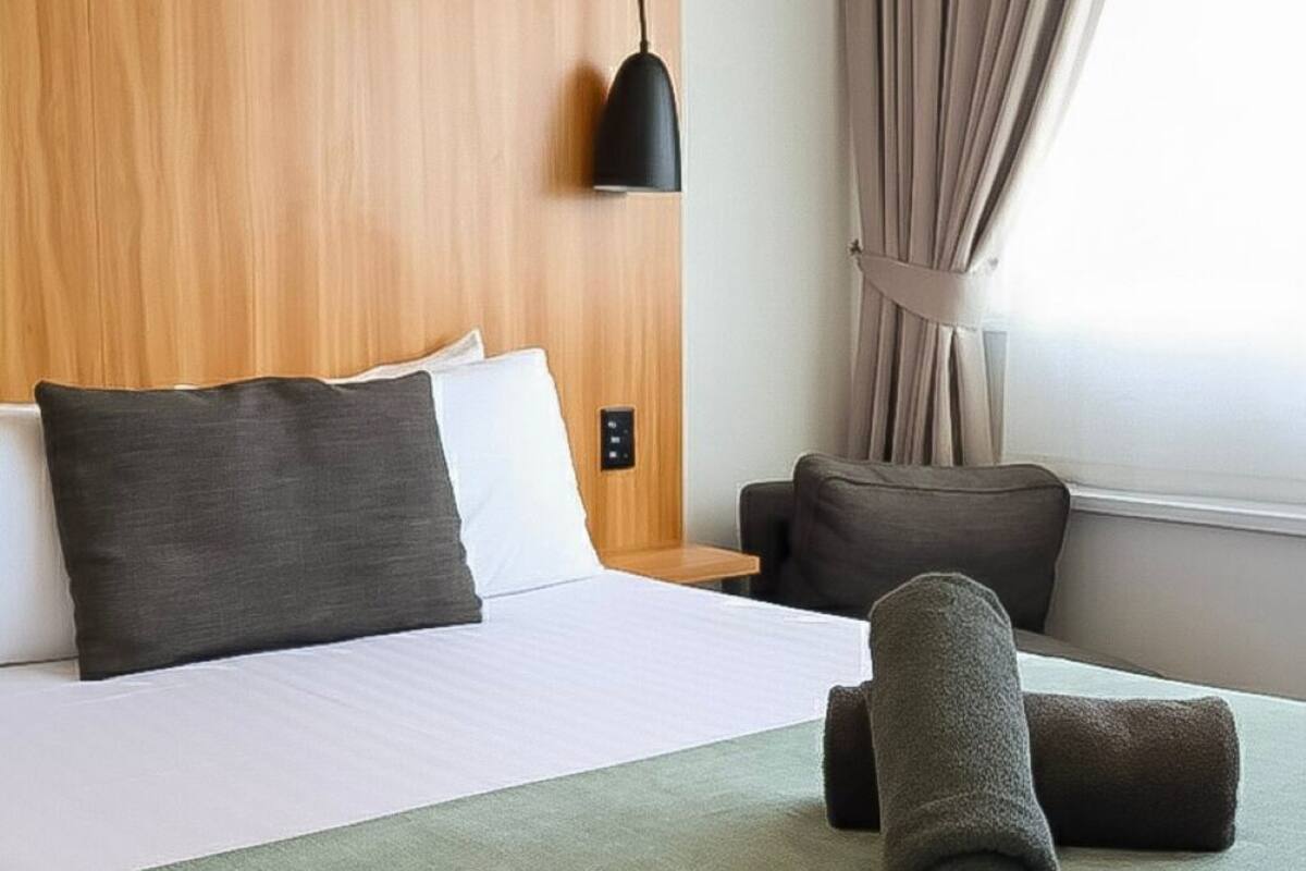 Ein Hotelzimmer mit einem hölzernen Kopfteil, einem Bett mit weißen und dunkelgrauen Kissen, einer grünen Tagesdecke, einem aufgerollten Handtuch, einer schwarzen Pendelleuchte, einem braunen Sofa und einem Fenster mit Vorhängen.
