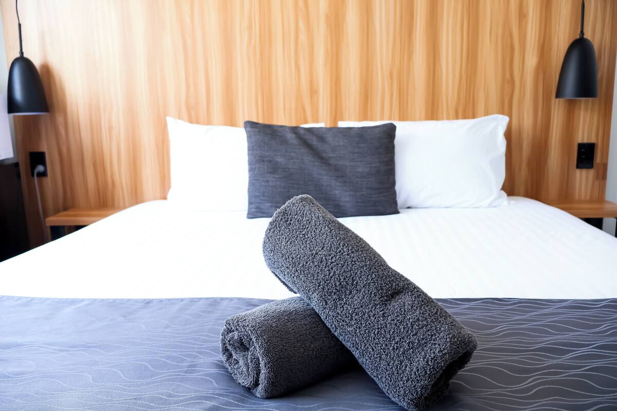 Ein ordentlich bezogenes Bett mit grauer Decke und Kissen, einem Holz-Kopfteil und zwei schwarzen Pendelleuchten.