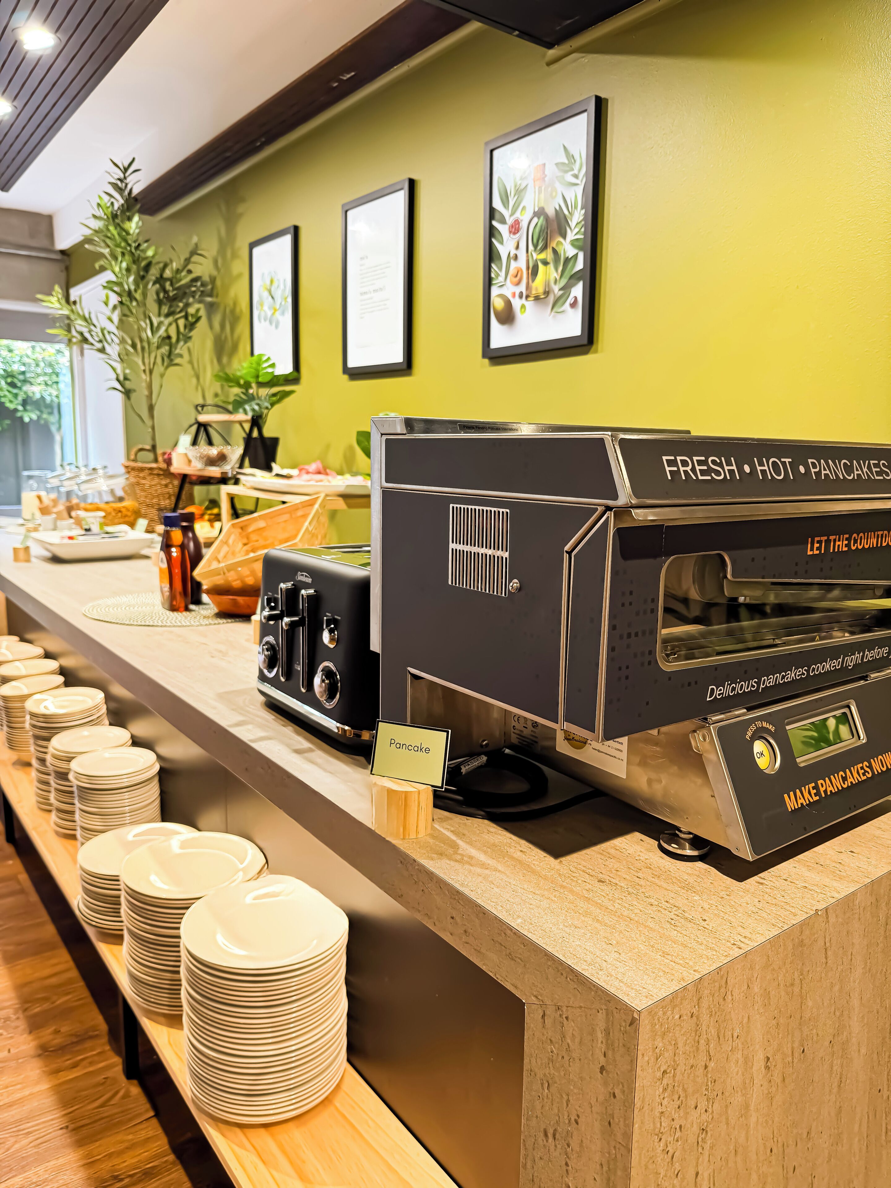 A breakfast buffet with a fresh hot pancakes station, a sign indicating the pancakes are made to order, and a variety of breakfast items displayed on the counter.