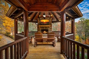 A wooden deck with a stone fireplace, two Adirondack chairs, and a view of autumn foliage.