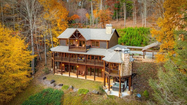 A large log cabin with multiple decks and a stone chimney, surrounded by autumn foliage.