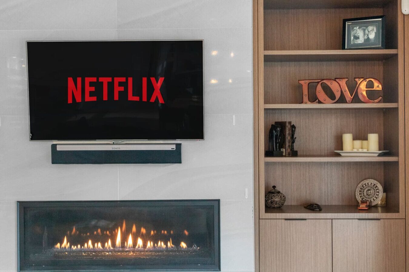 Ein flachbildfernseher an der Wand montiert, mit dem Wort "NETFLIX" in roter Schrift, darunter ein Kamin und ein Holzregal mit Dekoration und dem Wort "LOVE" in orangefarbener Schrift.