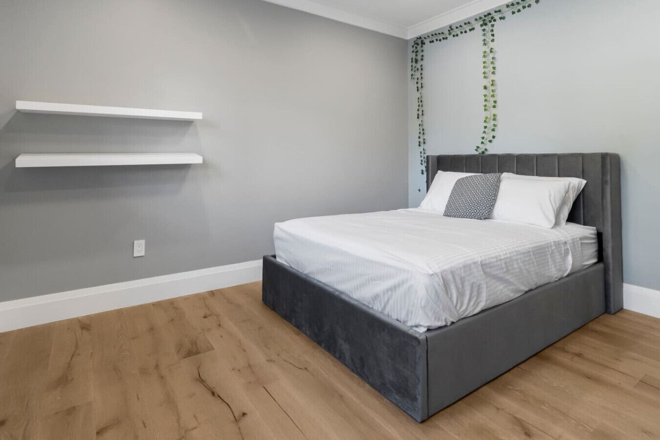 A bedroom with a grey bed, white bedding, a grey headboard, and a wooden floor. There are two white shelves on the wall and a power outlet below them.