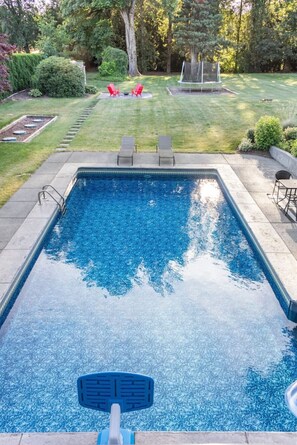 Una piscina rectangular con un trampolín, rodeada por un césped cuidado y algunas sillas rojas.