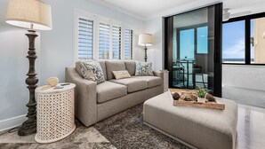 A living room with a beige sofa, two side tables, a glass door leading to a balcony, and a view of the ocean. - Beachside Bliss with Stunning Gulf Views & Resort Amenities – Lander's Sunset Lagoon - Roelens (Fort Myers Beach)
