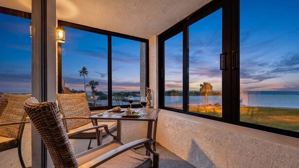A balcony with wicker chairs, a table with a bottle of wine, and a view of the ocean at dusk. - Beachside Bliss with Stunning Gulf Views & Resort Amenities – Lander's Sunset Lagoon - Roelens (Fort Myers Beach)