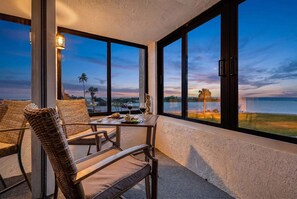 A balcony with wicker chairs, a table with a bottle of wine, and a view of the ocean at dusk.