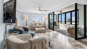 A spacious living room with a large flat-screen TV, a ceiling fan, and a glass door leading to an outdoor area. - Beachside Bliss with Stunning Gulf Views & Resort Amenities – Lander's Sunset Lagoon - Roelens (Fort Myers Beach)