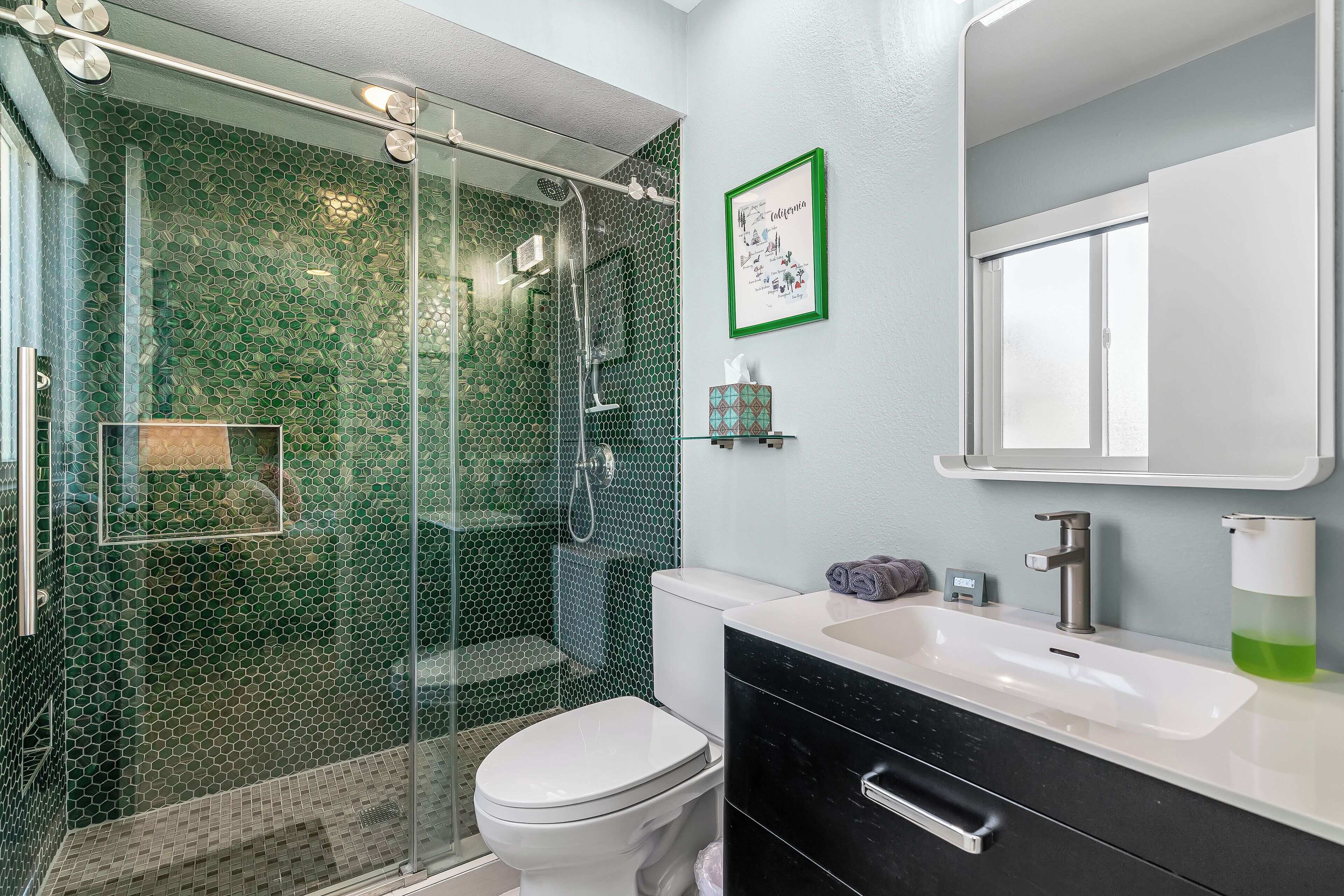 A bathroom with a glass-enclosed shower, a white toilet, a black sink with a mirror above it, and a framed picture on the wall.