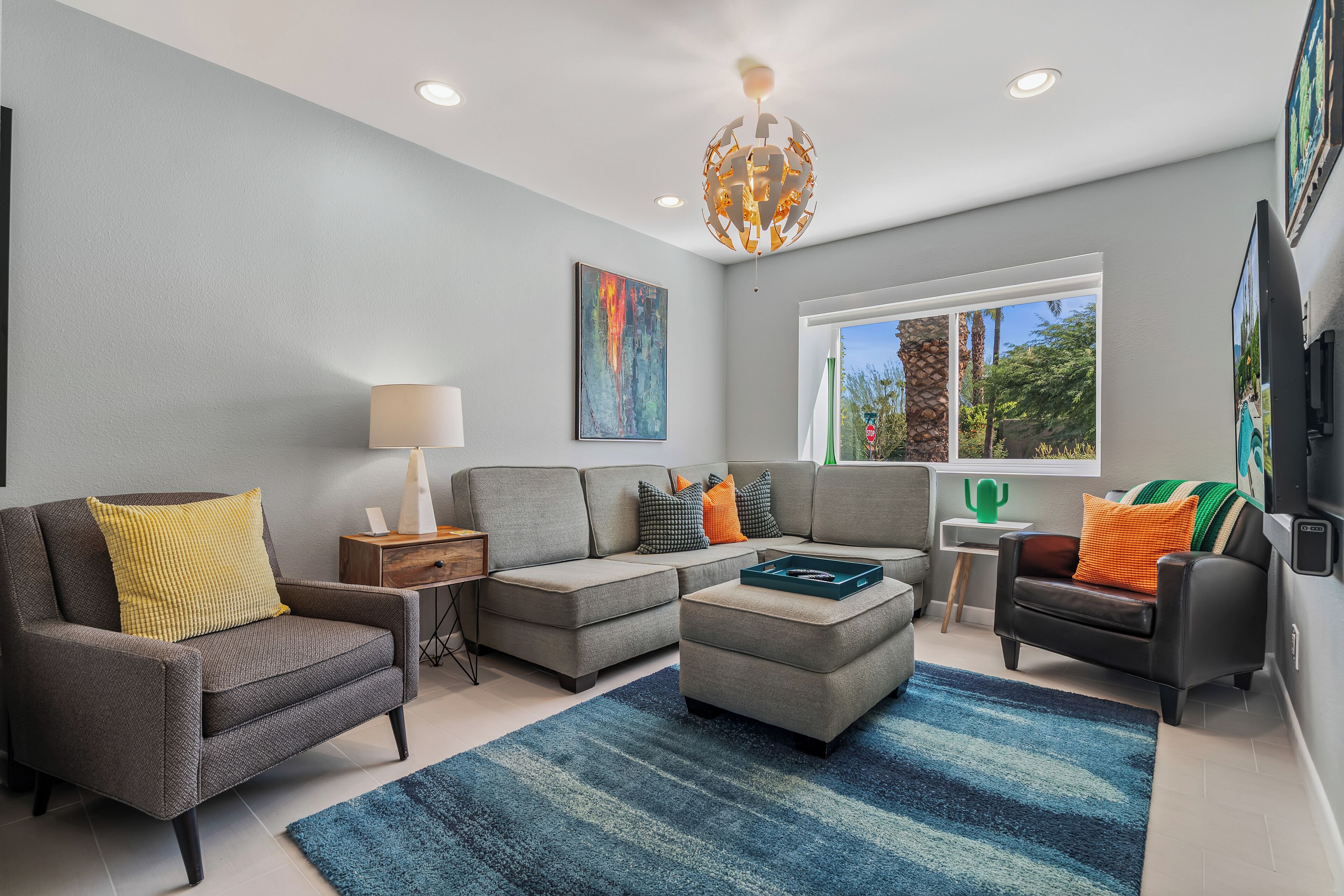 A modern living room with a grey sofa, a coffee table, and a TV. There is a window with a view of trees and a cactus.