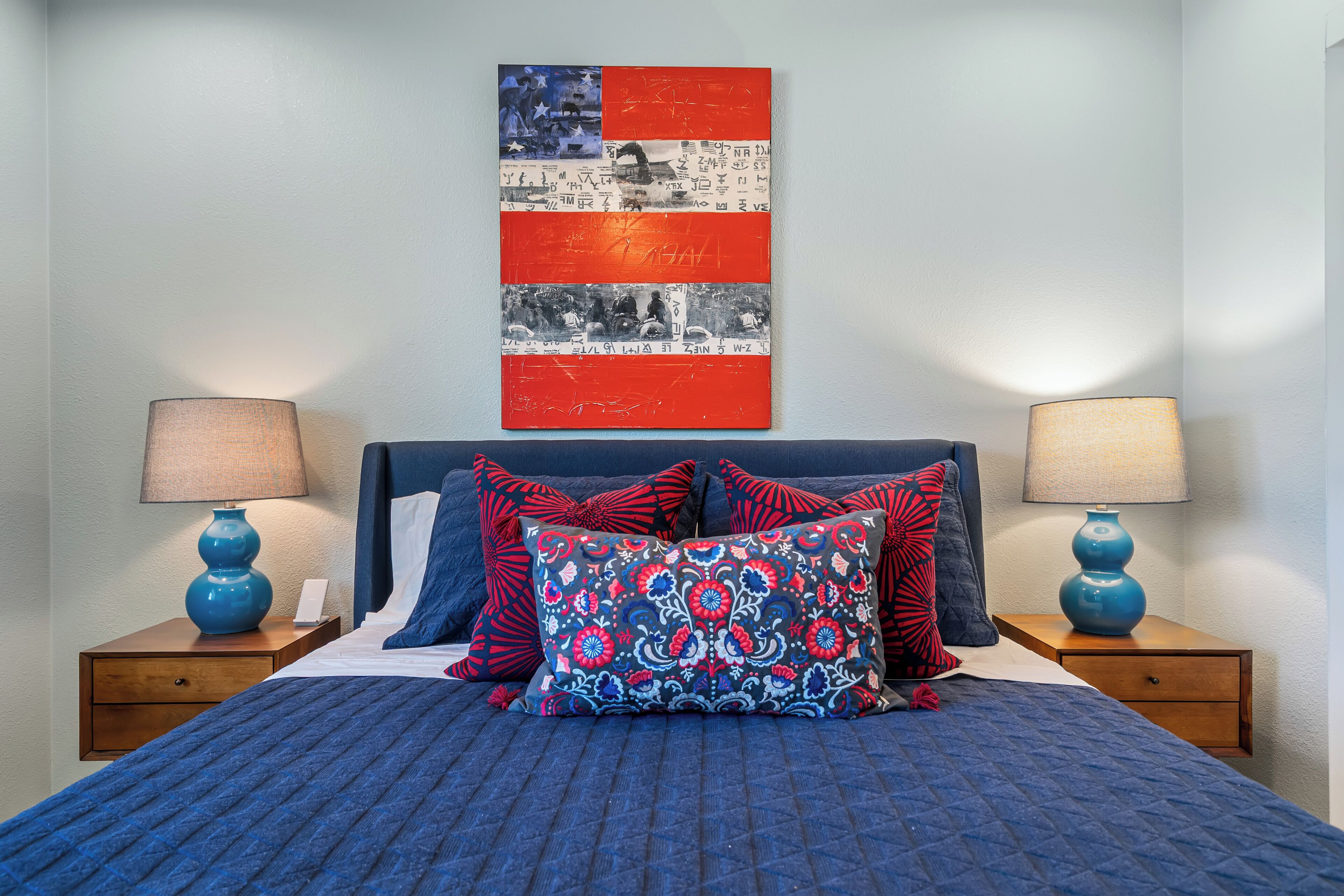 A neatly made bed with blue bedding and decorative pillows, flanked by two bedside tables with lamps, and a wall-mounted artwork above.