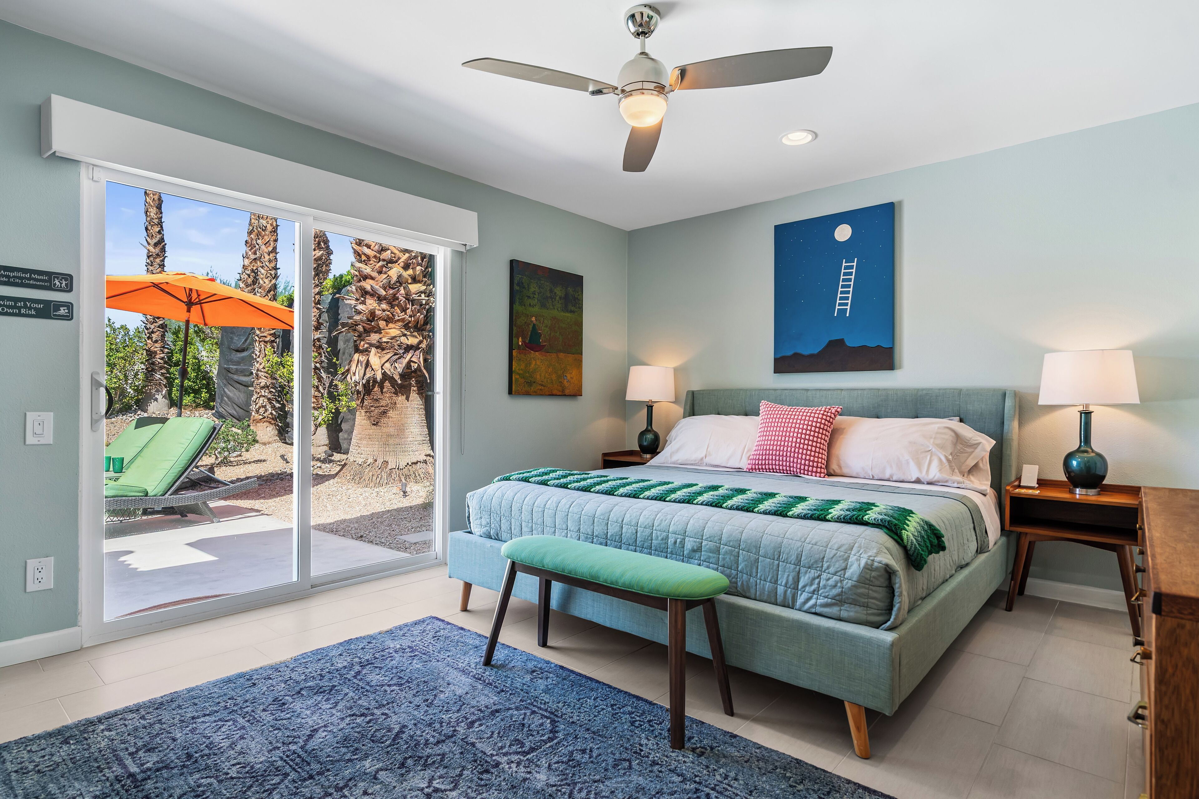A bedroom with a bed, two bedside tables, a ceiling fan, a nightstand, a bench, a large window, and a view of the outdoors.