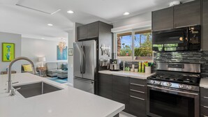 A modern kitchen with stainless steel appliances, a large island with a sink, and a view of the outdoors. - Desert Gem - Bold Design, Poolside Fun, Spa, BBQ & Views (Palm Springs)