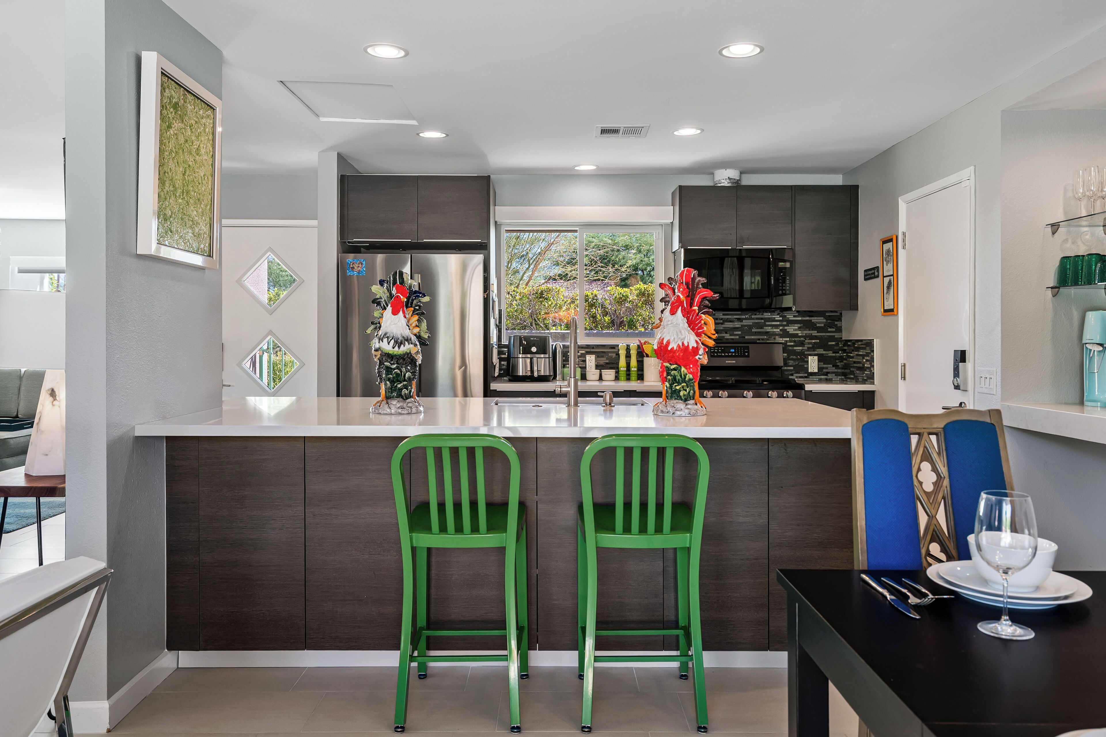 Una cocina moderna con una isla central, dos taburetes verdes, una mesa de comedor con un plato blanco y una copa, y vistas al exterior a través de una ventana.