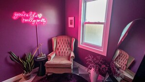 Ein Zimmer mit pinker Wand, einem Neonlichtschild, einem gepolsterten Sessel, einer Topfpflanze und einem Fenster.