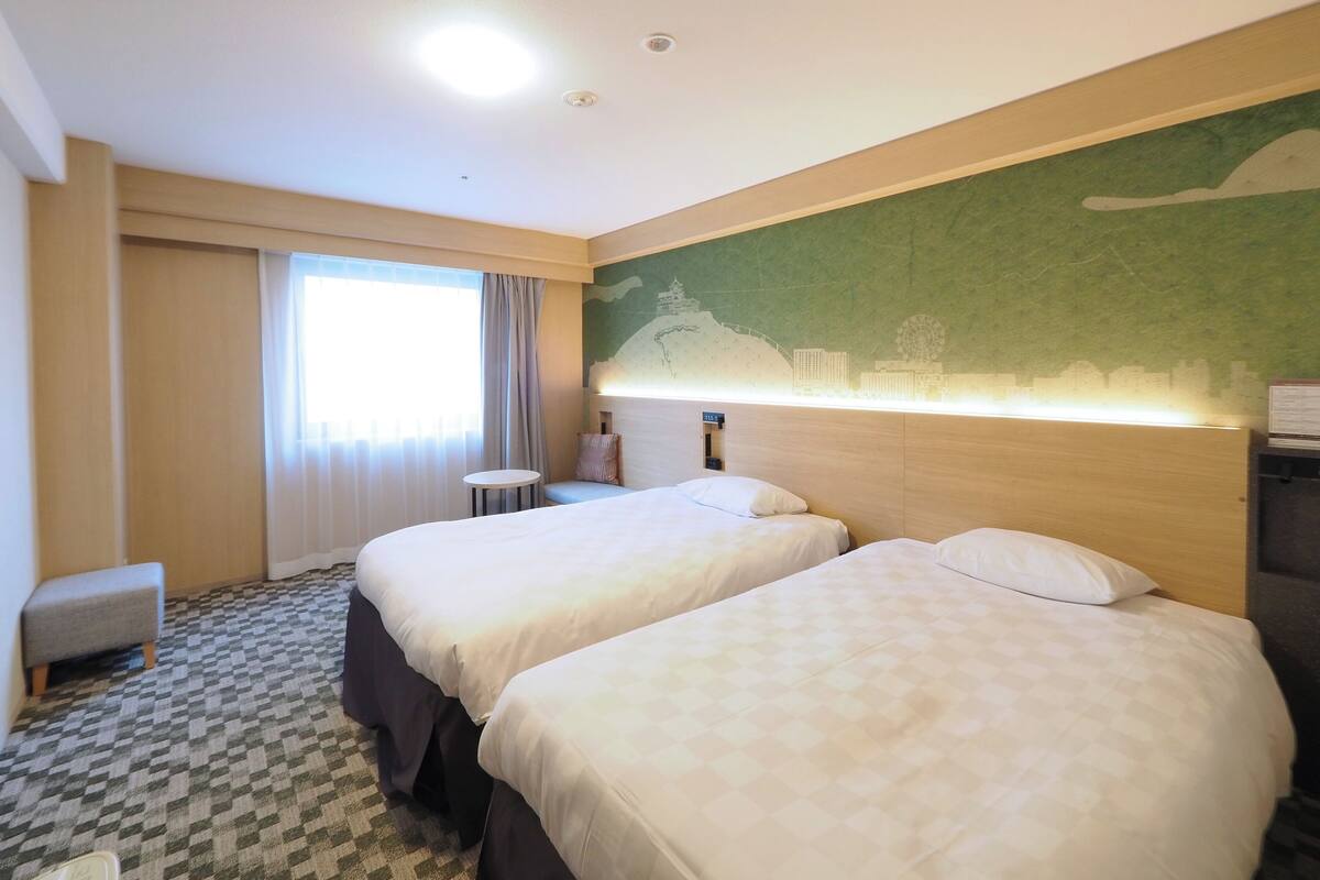 A hotel room with two beds, a small table, a bench, a window with curtains, and a wall mural.