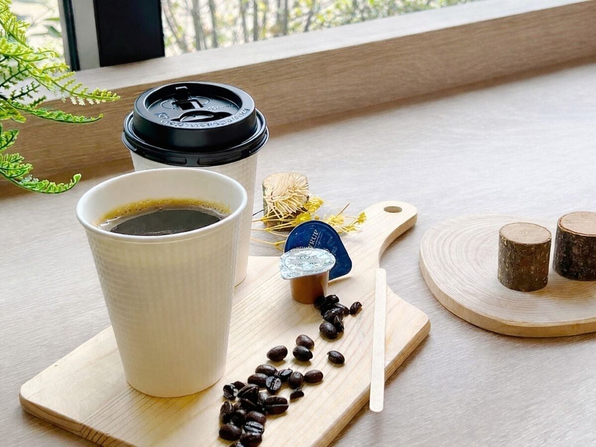 A cup of coffee on a wooden tray with coffee beans, a spoon, and a small container of creamer.