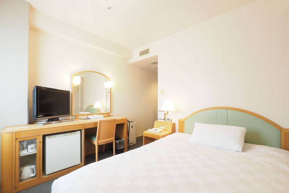 A hotel room with a bed, a desk with a computer, a television, a mirror, and a nightstand with a lamp.