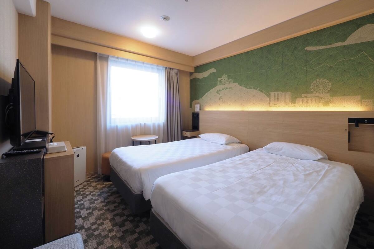 A hotel room with two beds, a desk with a computer, a TV, and a window with curtains.
