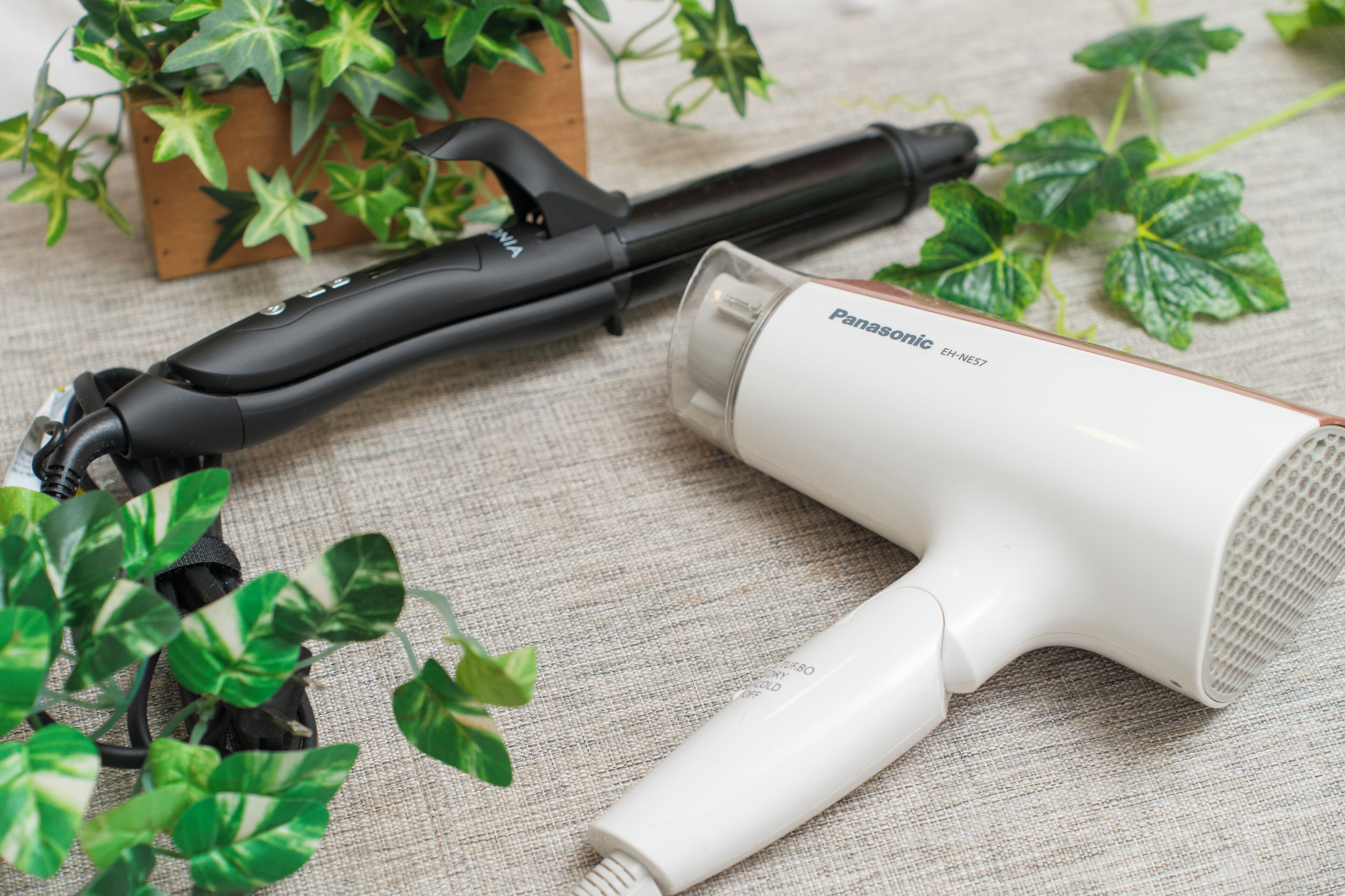 A Panasonic EH-NE57 hair dryer and a black hair styling tool on a textured fabric surface with green plants in the background.