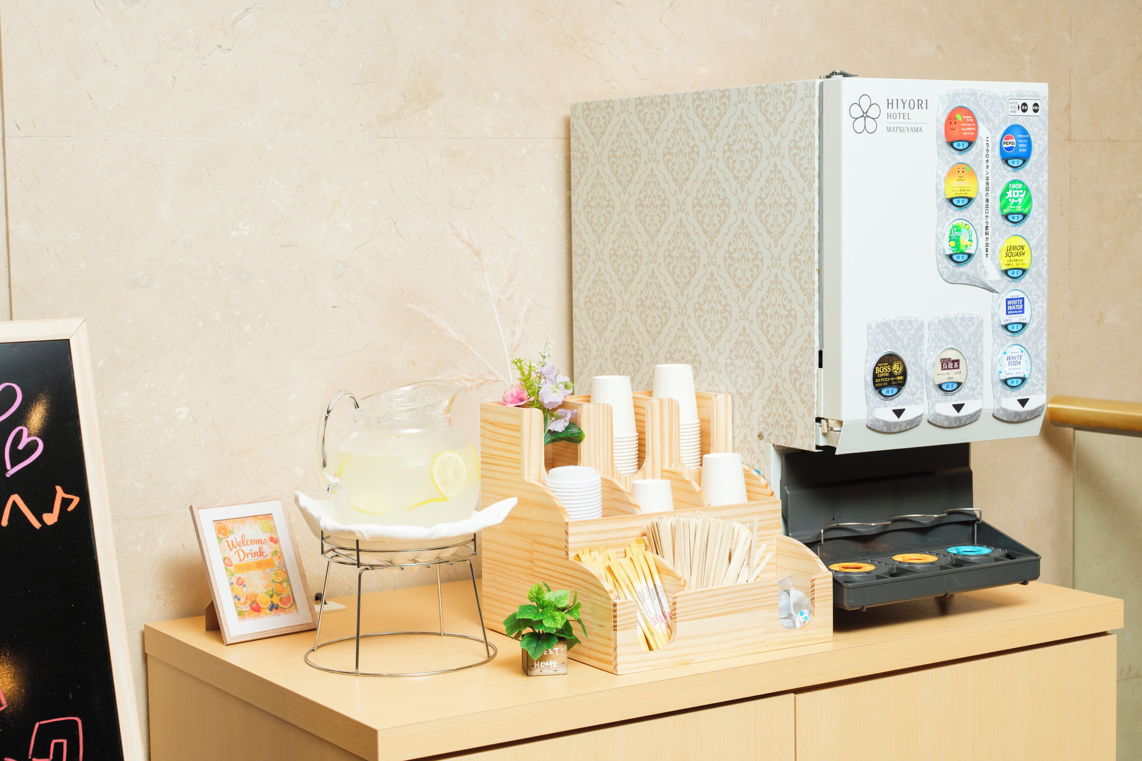 Dispensador de bebidas con diversas opciones, una jarra de limonada, vasos desechables y una planta pequeña sobre una encimera de madera.