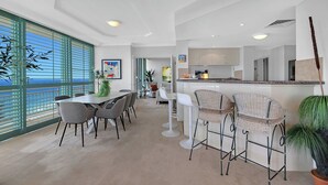 A modern kitchen with white cabinets, a central island, and a dining area with a table and chairs. - Panoramic Ocean View Sub-Penthouse 3mins to Beach (Surfers Paradise)