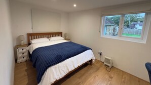 Une chambre avec un lit en bois, du linge de lit blanc, une table de chevet avec une lampe, une fenêtre donnant sur des maisons et un radiateur.